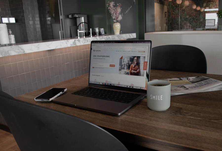 En dator i ett kök med en kaffekopp, mobiltelefon och tidning bredvid. På skärmen är det Avitus ITs kund Legalbuddy